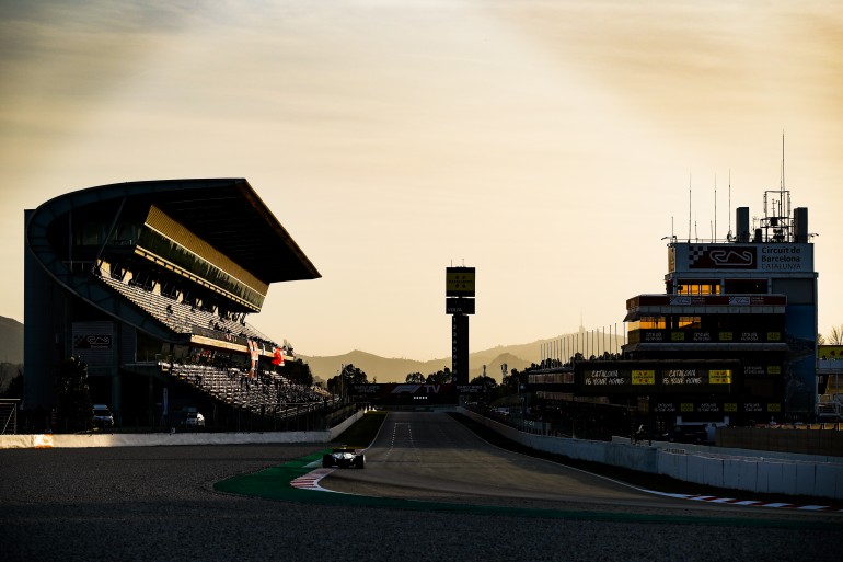 Circuito de Barcelona no fim de tarde com carro na tomada de curva durante os testes e arquibancada, boxes e torre ao fundo